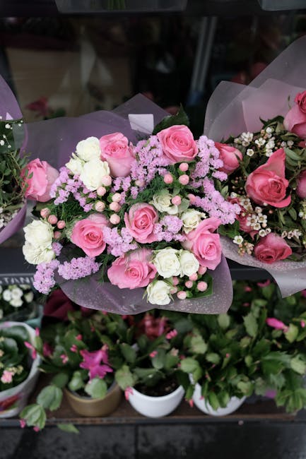 A floral bouquet featuring pink roses, white spray roses, and light pink astilbe arranged with green foliage. The flowers are wrapped in translucent purple tissue paper, with some greenery and small w