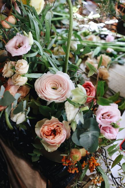 A close-up of a floral arrangement featuring soft pink and peach ranunculus, white and blush lisianthus, and sprigs of orange small flowers, all arranged with lush green foliage and eucalyptus leaves.