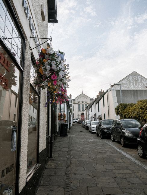 A hanging floral arrangement outside a shop along a narrow cobblestone street in Dalston, featuring a vibrant mix of white, pink, yellow, and purple flowers with lush green foliage, attached to a blac