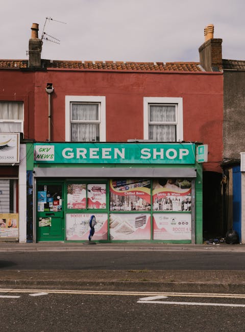The image features the facade of a small green shop with a prominent green sign reading 'GREEN SHOP' in white letters, situated on a street with a red brick building above. The shop's large glass wind