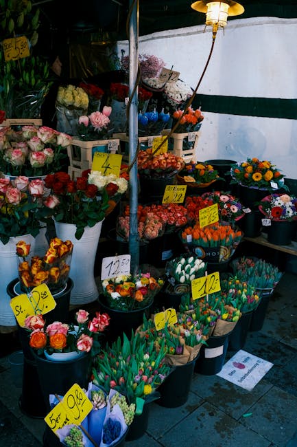A vibrant display of fresh flowers at a market stall, featuring a variety of floral arrangements in buckets and baskets. Prominent flowers include pink and cream roses, orange and yellow sunflowers, a