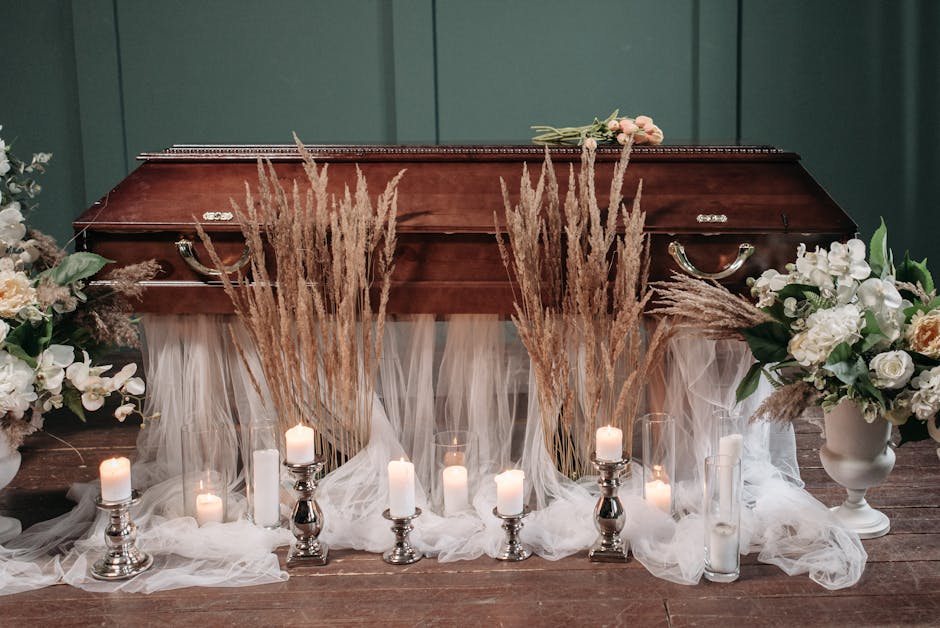 A floral arrangement featuring white and cream roses, peonies, and greenery in white ceramic vases, complemented by tall dried pampas grass stems. The flowers are set on a dark wooden surface, with a 