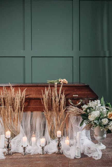 A floral tribute placed on a polished wooden casket, crafted with soft pink roses, white peonies, and greenery, surrounded by tall, golden wheat stalks and illuminated by small, elegant candles in sil