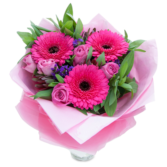 I picture this bouquet as an interplay of confident colour and soft romance: three vivid fuchsia Gerbera daisies open wide, their petal rays creating a graphic centrepiece against the softer forms around them. Close to those gerberas, roses in a dusty lavender-pink unfurl with composed elegance, their tight buds signalling freshness and a gentle perfume. Dotted throughout are clusters of purple statice that offer a papery, meadow-like texture and a cool visual punctuation against the warm pinks. Supporting foliage-broad salal leaves with the occasional rounded sprig of eucalyptus-builds a verdant backdrop that makes the colours sing and adds a tactile crispness to the bouquet. The flowers are wrapped in an inner sheet of soft matte pale-pink tissue, then swathed in a layer of translucent, slightly crinkled fuchsia-pink cellophane that gives the whole presentation a subtle glow. The lower portion reveals the top rim of a clear glass vase, suggesting both readiness for display and the clean lines of contemporary table styling. Under soft diffused light the petals pick up a velvety sheen and the aroma is light and approachable-roses giving a gentle scent while the gerberas offer a fresh, green hint. This arrangement feels at home for celebrations big and small: birthdays, Mother's Day, anniversaries, or as a thoughtful surprise after a walk through Ridley Road Market or a picnic by the Eastern Curve Garden. Made with the care of a local florist, it's crafted to brighten interiors across Dalston and nearby Hackney.